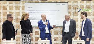 Vier Männer und eine Frau mit Mikrofonen sprechen auf einer Bühne vor einer Holzwand an der ein großes Display hängt.