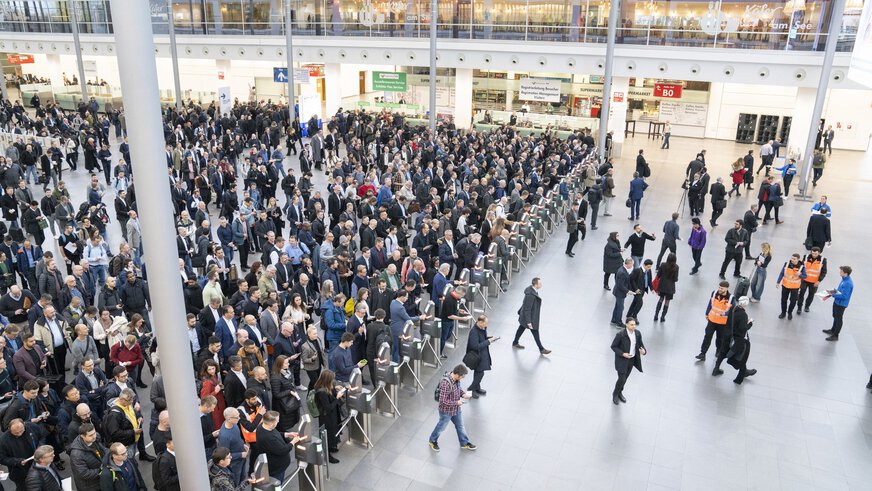 Blick von der Seite über die Eingangshalle der productronica Messe mit zahlreichen wartenden Besuchern an der Ticketkontrolle. Einige Besucher sind schon hindurch.