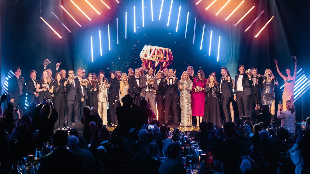 Eine große Gruppe in formeller Kleidung feiert auf der Bühne unter hellen Lichtern bei einer Preisverleihung.