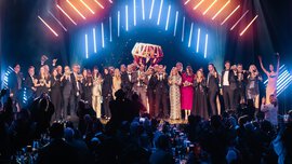 Eine große Gruppe in formeller Kleidung feiert auf der Bühne unter hellen Lichtern bei einer Preisverleihung.