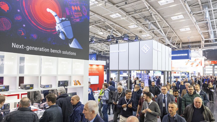 Zahlreiche Besucher gehen an den Ausstellerständen auf der productronica entlang. Im Fokus liegen die Werbetafeln mit den Ausstellerlogos unter der Hallendecke.