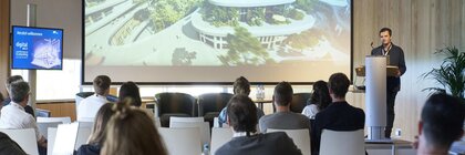 Presentation on a stage with a speaker at a podium, seated audience and a large architectural design on the screen.