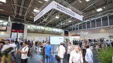 People walk through an exhibition hall with stands and a cordoned-off demonstration area.