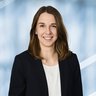 Eine lächelnde Frau mit langen braunen Haaren, schwarzem Jackett und weißer Bluse.