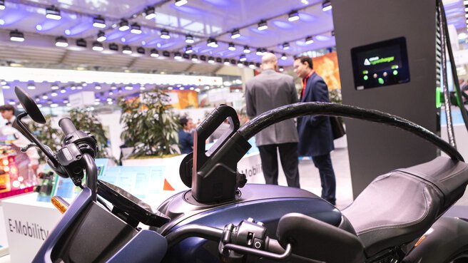 Electric motorcycle on display at an exhibition stand with people in the background and modern lighting above.