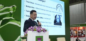 A man in a suit speaks on a podium during a forum, behind him a presentation slide about challenges is shown.