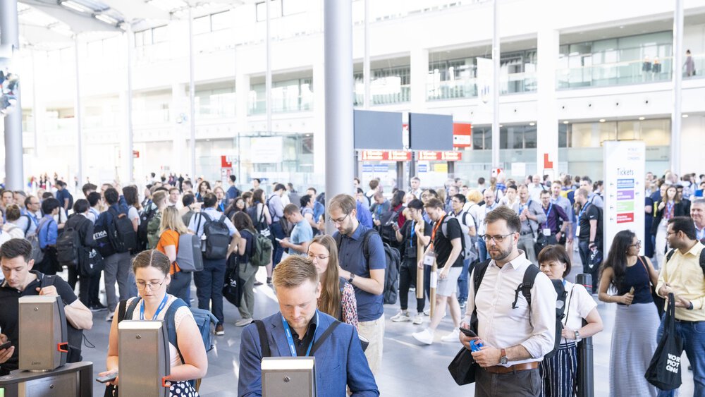 A large group of people stand in front of the ticket barriers in the exhibition entrance hall.