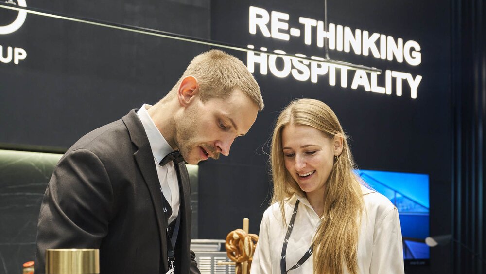 Ein Mann und eine Frau im Gespräch an einem Messestand auf der EXPO REAL in München. Der Mann in Anzug und Fliege schreibt etwas auf ein Blatt Papier, während die Frau in weißer Bluse lächelnd danebensteht. Im Hintergrund hängt ein Schild mit der Aufschrift „RE-THINKING HOSPITALITY“ vor einer modernen, dunkel gehaltenen Standkulisse.