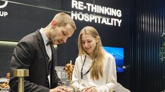 A man and a woman are talking at an exhibition booth at EXPO REAL in Munich. The man, wearing a suit and bow tie, is writing on a piece of paper while the woman in a white blouse smiles beside him. In the background, a sign reads “RE-THINKING HOSPITALITY” against a modern, dark-colored booth design.