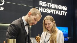 Ein Mann und eine Frau im Gespräch an einem Messestand auf der EXPO REAL in München. Der Mann in Anzug und Fliege schreibt etwas auf ein Blatt Papier, während die Frau in weißer Bluse lächelnd danebensteht. Im Hintergrund hängt ein Schild mit der Aufschrift „RE-THINKING HOSPITALITY“ vor einer modernen, dunkel gehaltenen Standkulisse.
