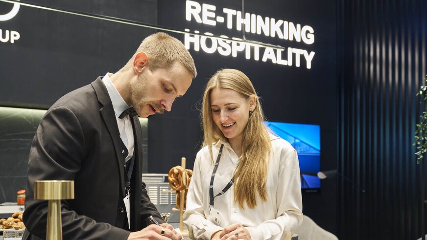 A man and a woman are talking at an exhibition booth at EXPO REAL in Munich. The man, wearing a suit and bow tie, is writing on a piece of paper while the woman in a white blouse smiles beside him. In the background, a sign reads “RE-THINKING HOSPITALITY” against a modern, dark-colored booth design.