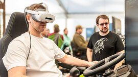 Eine Person, die ein VR-Headset trägt, sitzt in einem Simulatorstuhl und hält einen Joystick in der Hand, während ein Mann neben einem Computermonitor zusieht.
