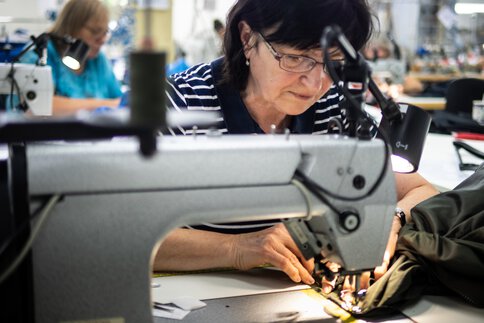 Eine Frau mit Brille arbeitet konzentriert an einer Nähmaschine. Im Hintergrund ist eine weitere Person bei der Arbeit zu sehen.