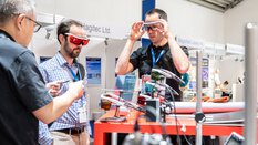 Two people try on modern safety goggles at an exhibition stand.