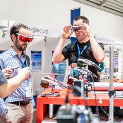 Two people try on modern safety goggles at an exhibition stand.