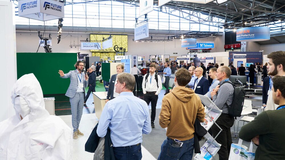 Mehrere Messebesucher stehen in einer Halle der productronica an einem Stand und hören einem Mann in kariertem Anzug zu, der in ein Mikrofon spricht.