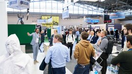 Mehrere Messebesucher stehen in einer Halle der productronica an einem Stand und hören einem Mann in kariertem Anzug zu, der in ein Mikrofon spricht.