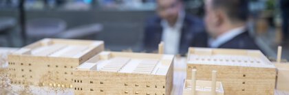 Architectural model of a building complex made of light wood with several rectangular structures, in the blurred background two people in conversation.