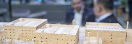 Architectural model of a building complex made of light wood with several rectangular structures, in the blurred background two people in conversation.