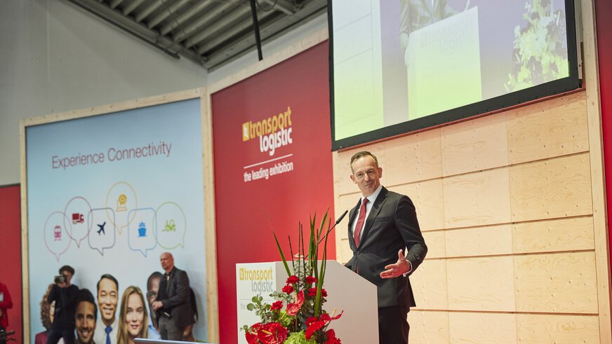 Ein Mann im Anzug und mit roter Krawatte spricht an einem Podium auf der Bühne, im Hintergrund eine Leinwand und mehrere Logos zur transport logistic Veranstaltung.