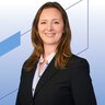 A woman with long brown hair wearing a black blazer is smiling against a blue geometric background.
