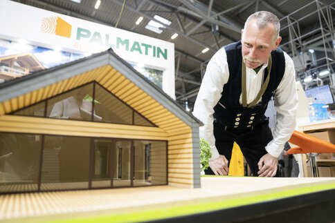 Während der BAU-Messe beugt sich ein Mann in Kluft zu einem Modellhaus aus Holz und Glas hinunter.