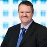 A smiling man with short dark hair, glasses and a goatee is wearing a shirt with a blue tie and a black jacket.