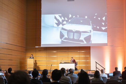 The audience in a modern conference hall watches a presentation on a large screen showing a car.