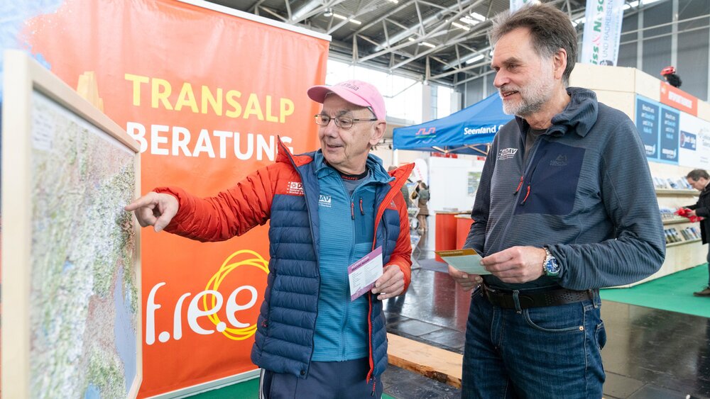 Zwei Männer betrachten und diskutieren auf einer Indoor-Veranstaltung mit Reise- und Abenteueranzeigen im Hintergrund eine große Karte.
