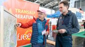 Zwei Männer betrachten und diskutieren auf einer Indoor-Veranstaltung mit Reise- und Abenteueranzeigen im Hintergrund eine große Karte.