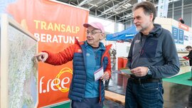 Zwei Männer betrachten und diskutieren auf einer Indoor-Veranstaltung mit Reise- und Abenteueranzeigen im Hintergrund eine große Karte.