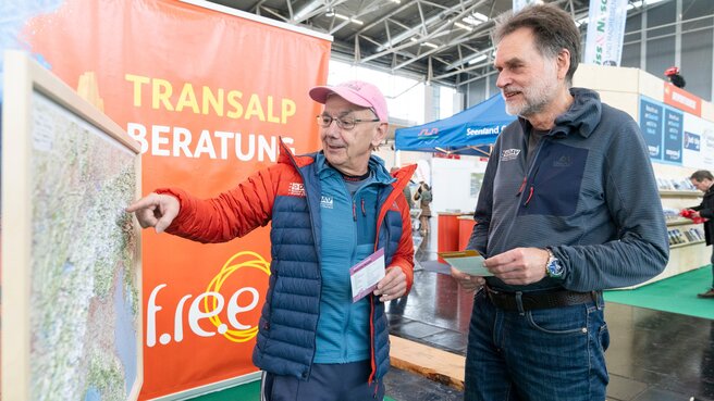 Zwei Männer betrachten und diskutieren auf einer Indoor-Veranstaltung mit Reise- und Abenteueranzeigen im Hintergrund eine große Karte.