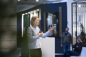 Eine Frau mit Brille, Ansteckmikrofon und einer Fernbedienung in der Hand hält einen Vortrag vor Publikum.