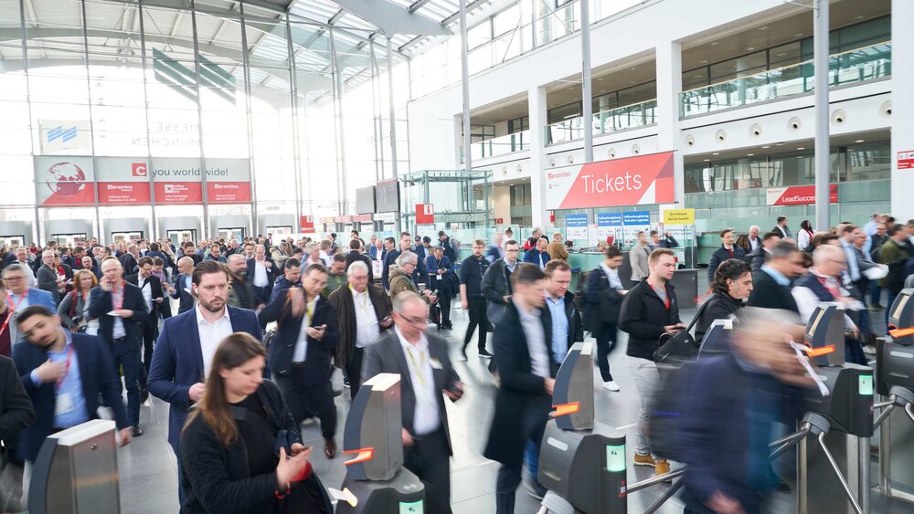 Eine Eingangshalle zur ceramitec mit zahlreichen Messebesuchern, die sich auf die Ticketkontrollen zubewegen. Im Hintergrund erkennt man einen Ticketverkaufsschalter.