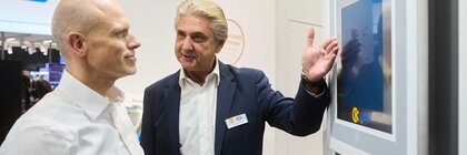 Zwei Männer im Business-Outfit diskutieren an einem Touchscreen während einer Elektronikmesse und betrachten digitale Präsentationsinhalte.