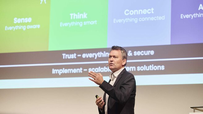A man speaks at a conference, with colorful infographic slides on a screen in the background.