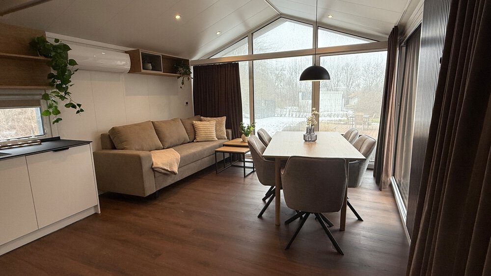 Ein Wohnzimmer mit Sofa, Essecke, großen Fensterfronten mit Vorhängen und einer Klimaanlage an der Wand.