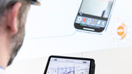 Eine Person mit Schutzhelm verwendet ein Tablet, das einen Gebäudegrundriss anzeigt, während im Hintergrund ein Smartphone zu sehen ist.