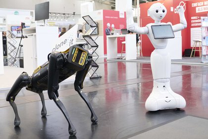 Two robots, one stands on four legs, the other stands upright, has a display and a face.