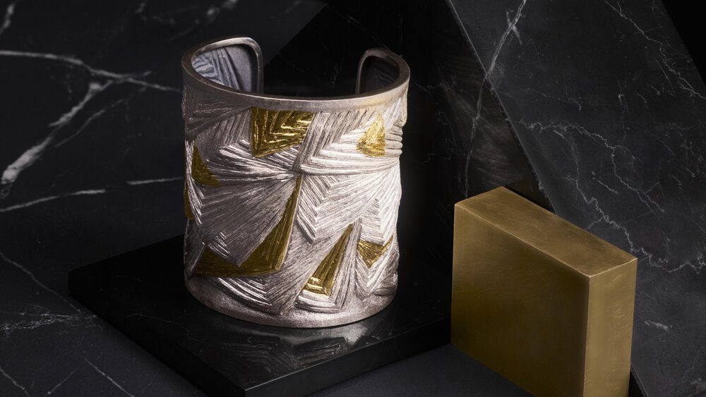 Silver and gold cuff with geometric pattern on a black marble surface with a rectangular brass block next to it.