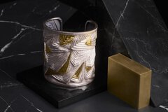 Silberne und goldene Manschette mit geometrischem Muster auf einer schwarzen Marmoroberfläche mit einem rechteckigen Messingblock daneben.