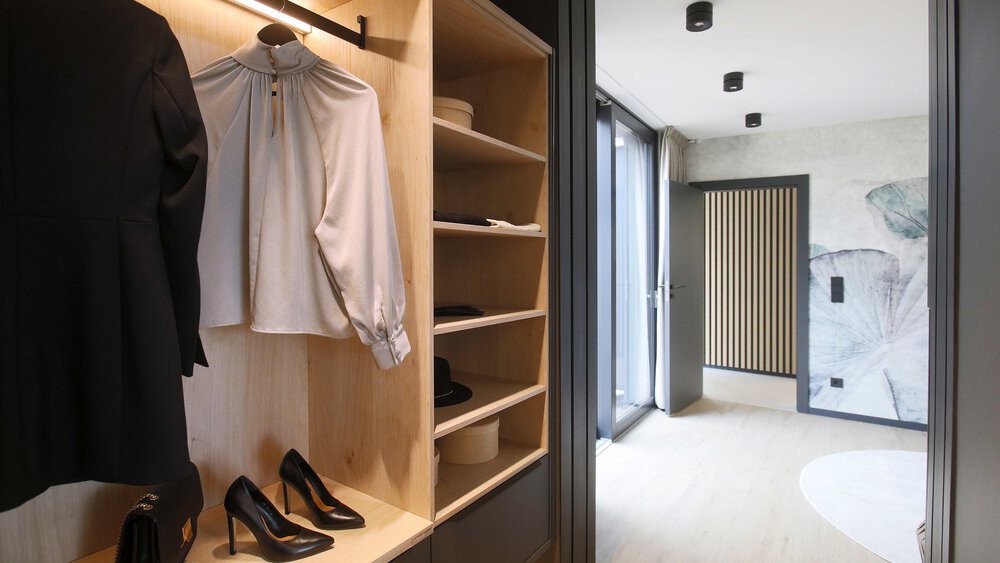 Offener Schrank mit Kleidung und Schuhen neben einer Tür, die zu einem gut beleuchteten Flur mit Wandkunst führt.