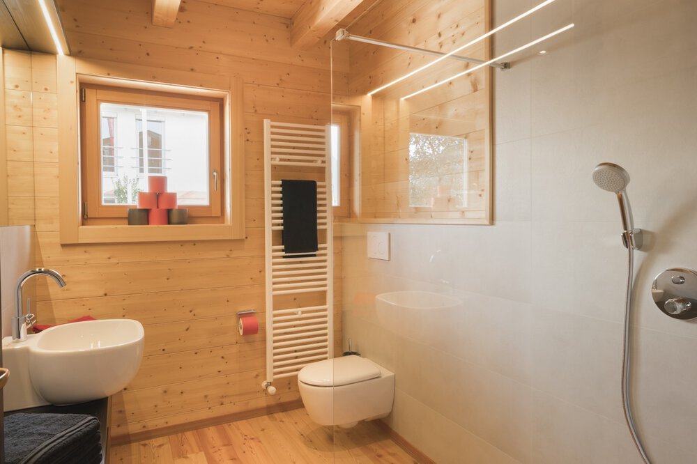 Holzbadezimmer mit Dusche, Wandwaschbecken, WC, Handtuchheizkörper und kleinem Fenster mit roten Kerzen.