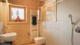 Holzbadezimmer mit Dusche, Wandwaschbecken, WC, Handtuchheizkörper und kleinem Fenster mit roten Kerzen.