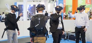 Vier Personen mit VR-Headsets und Rucksäcken interagieren in einer technischen Ausstellung mit blauem Bodenbelag.