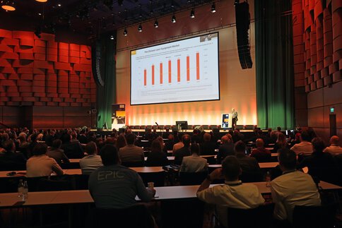 Vortrag auf einer Fachkonferenz vor großem Publikum mit Präsentation einer Marktanalyse auf einer Großbildleinwand.