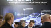 Besucher der productronica stehen vor einem Ausstellerstand. An der Wand sieht man ein großes futuristisches Bild eines Autos mit AI-Steuerung.