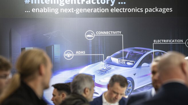 Besucher der productronica stehen vor einem Ausstellerstand. An der Wand sieht man ein großes futuristisches Bild eines Autos mit AI-Steuerung.