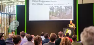 A man gives a presentation to an audience at INTERFORST. A screen with a presentation slide can be seen behind him.