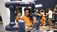 Two people are working at a trade fair stand on a laptop in front of which a robot arm can be seen.